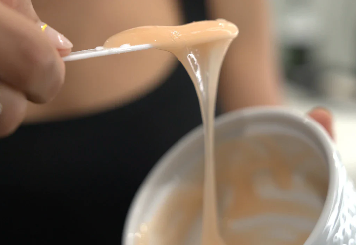 Close-up of a hand holding a spoon with creamy substance over a container.