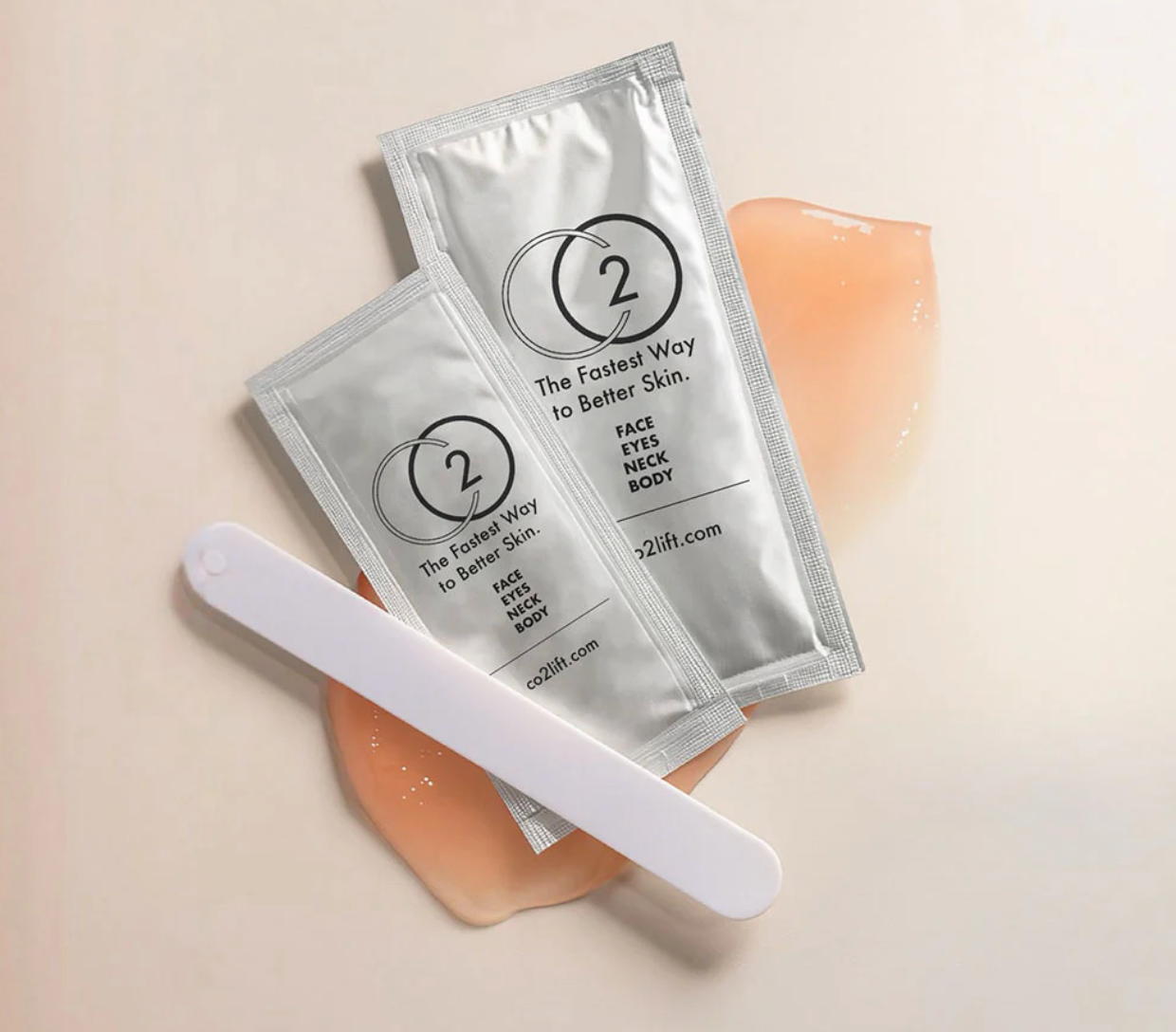 Two silver packets with a logo and text, a white spatula, and a beige cream on a beige background.