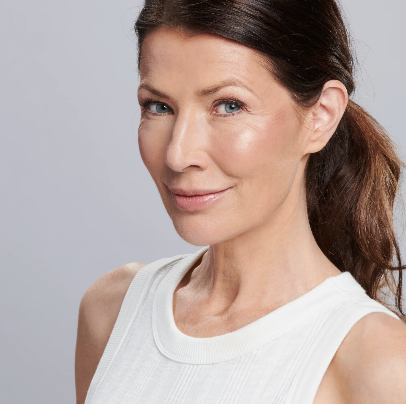Woman with a ponytail wearing a white tank top against a gray background